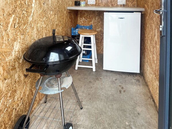 A compact kitchen with a black grill, a white refrigerator, and a stool.
