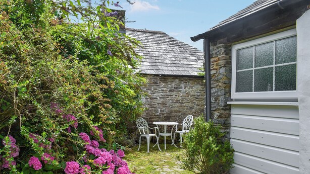 A stone building with a tiled roof, a white window with a textured glass pane, and a small garden area with pink flowers and a potted plant. - 1 bedroom accommodation in Camelford (Camelford)