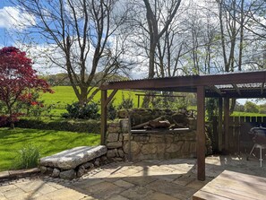 A stone patio with a pergola, a barbecue grill, and a view of a green field and trees.