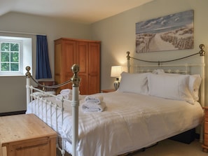 Una cama bien hecha con sábanas blancas, una mesita de noche de madera, un armario de madera, un cuadro en la pared y una ventana con cortinas.