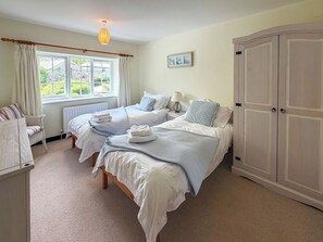 A bedroom with two beds, a window with curtains, a radiator, a bedside table with a lamp, a chair, and a wardrobe.