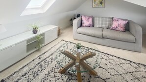 A loft-style room with a skylight, a grey sofa with floral cushions, a white console unit, and a glass coffee table.