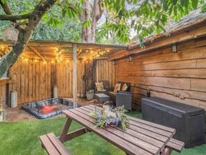 Een houten terras met een jacuzzi, rieten meubels en een picknicktafel met bloemen.