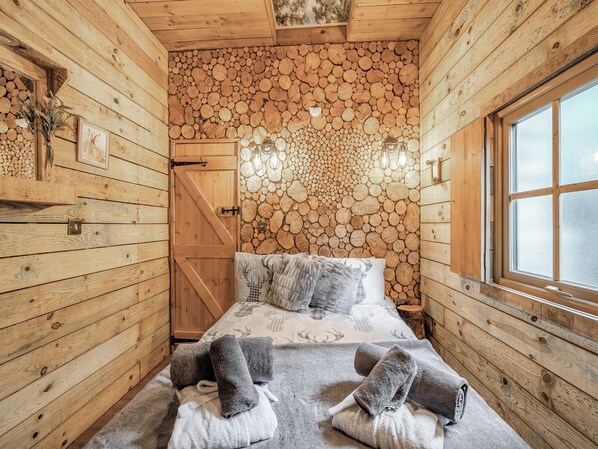 Ein Zimmer in einer Holzhütte mit einem Bett, Handtüchern, einer Holzwand mit Baumstämmen, einem Fenster und einer Tür.
