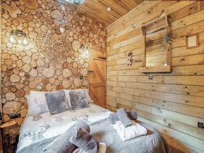 Interior de una cabaña de madera con una cama, una pared de troncos apilados, una puerta de madera y un espejo.