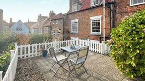 A patio with a table and chairs, surrounded by a white picket fence, and brick houses in the background.