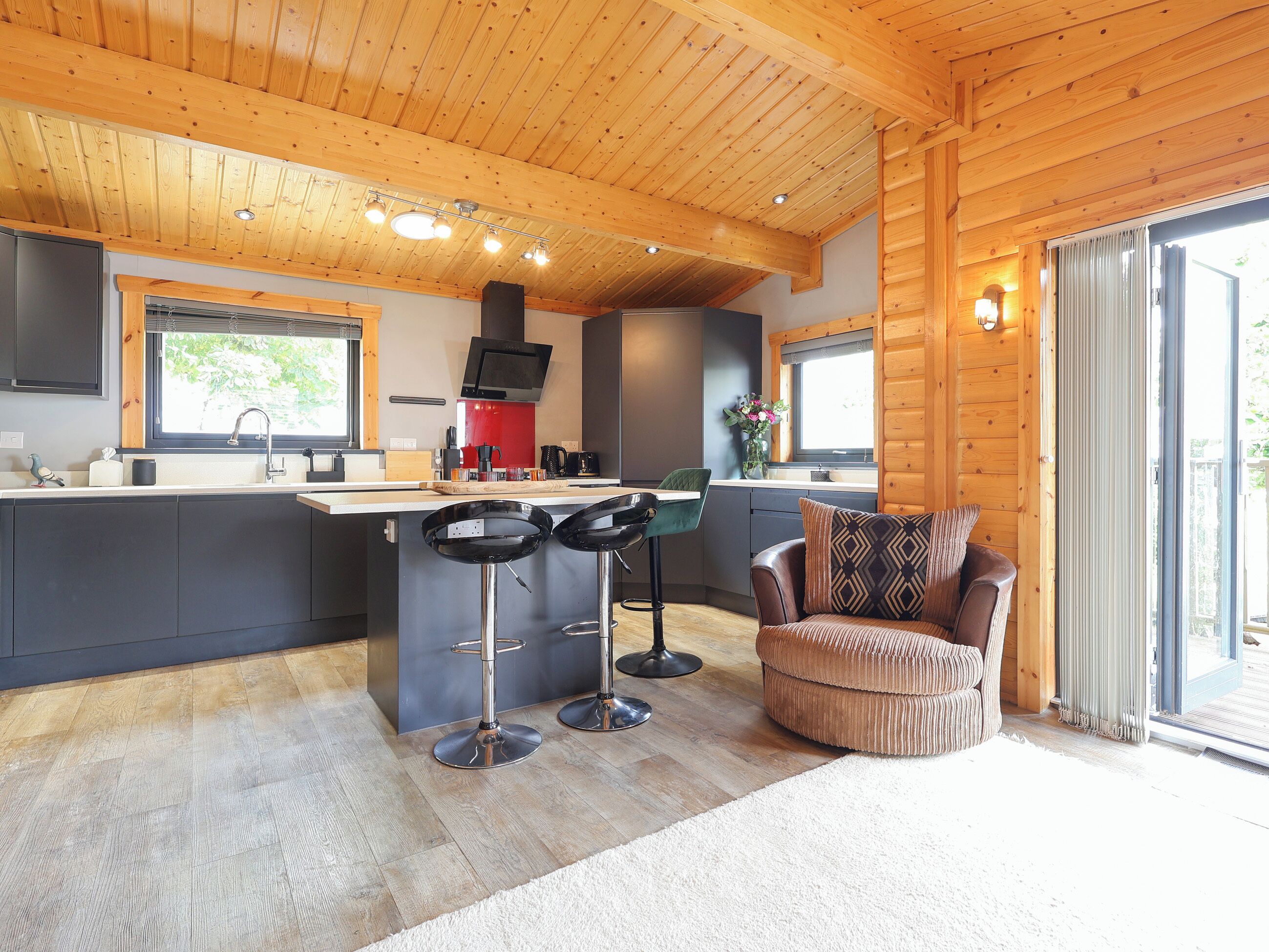 A modern kitchen with a central island, bar stools, a sofa, and a sliding glass door leading to an outdoor area.