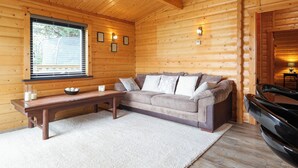 A cozy cabin interior with a wooden ceiling, a sofa with cushions, a coffee table, a window with blinds, and a car parked outside.