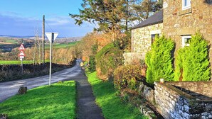 Une étroite route de campagne, avec une maison en pierre sur la droite, un chemin piétonnier et un ciel dégagé.