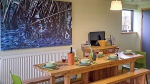 Un coin repas avec une table en bois dressée pour quatre personnes, des chaises vertes, un grand tableau au mur, une fenêtre donnant sur un paysage et un radiateur placé sous la table.