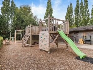 A wooden play structure with a slide, climbing wall, and stairs, surrounded by trees and a gravel ground.