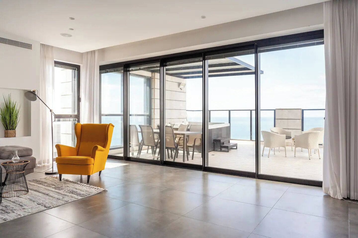 A modern living room with a yellow armchair, a dining area with white chairs, and a balcony with a view of the sea.