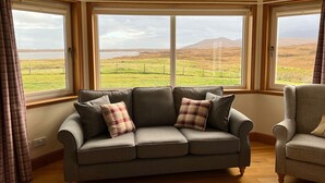 A room with a grey sofa and armchair, large windows with a view of a grassy landscape and mountains. - Airigh nam Fiadh (South Uist)