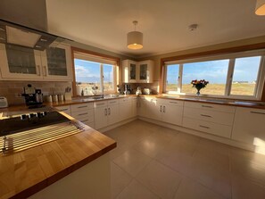 A modern kitchen with white cabinets, a wooden countertop, and large windows offering a view of the countryside. - Airigh nam Fiadh (South Uist)