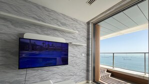 A modern hotel room with a large TV mounted on a marble wall, a sliding glass door leading to a balcony with a view of the sea, and a built-in white entertainment unit. - Flex Ottomare Suites (İstanbul)