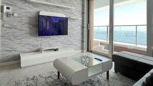 A modern living room with a marble wall, a flat-screen TV, a glass coffee table, and a large sliding glass door leading to a balcony with a view of the sea. - Flex Ottomare Suites (İstanbul)