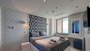 A modern bedroom with a patterned wallpaper, a large bed with pillows, a bedside table, a mirror, and a window with a view. - Flex Ottomare Suites (İstanbul)