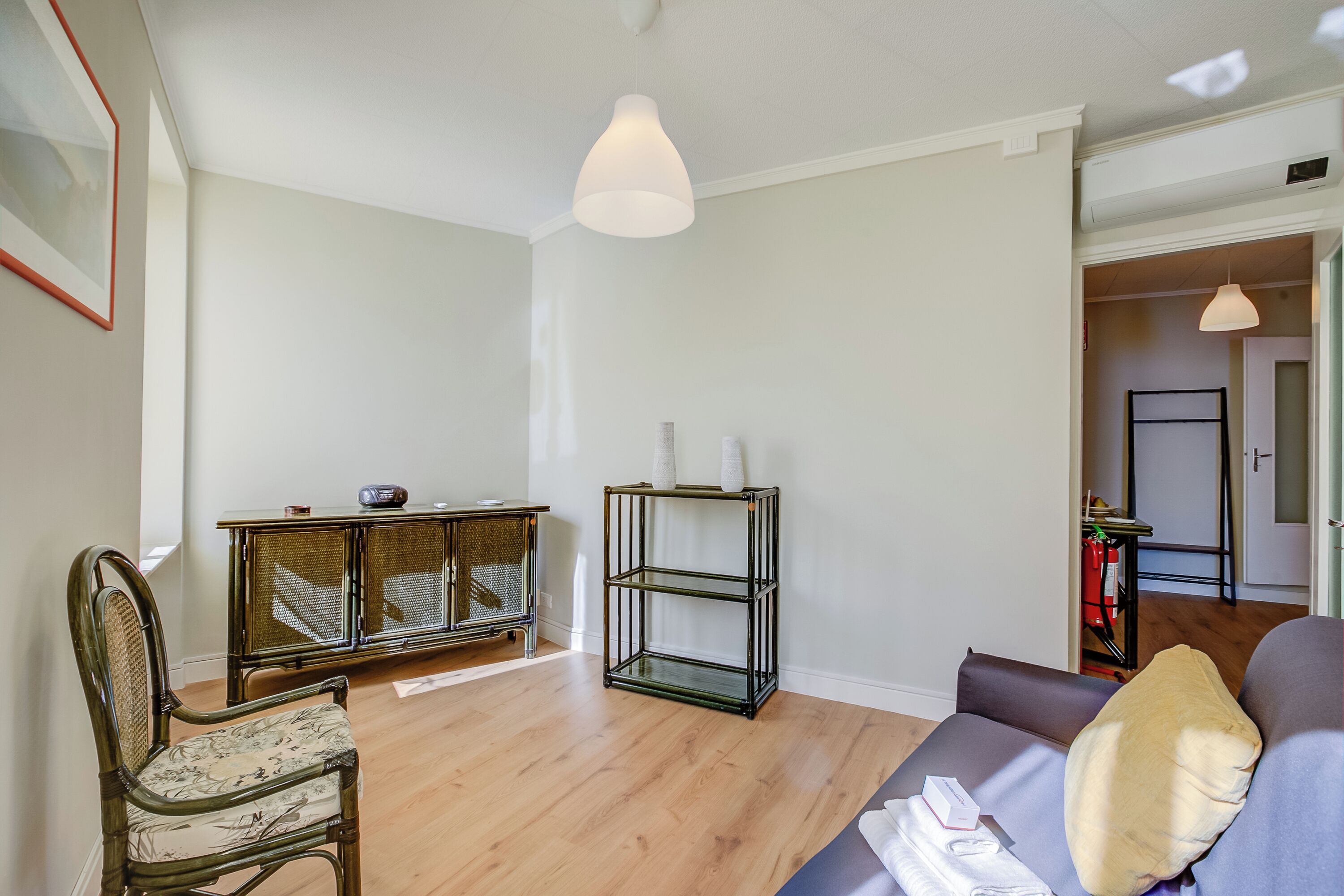 A living room with a wooden floor, a sofa, a chair, a glass shelving unit, and a hanging light fixture.