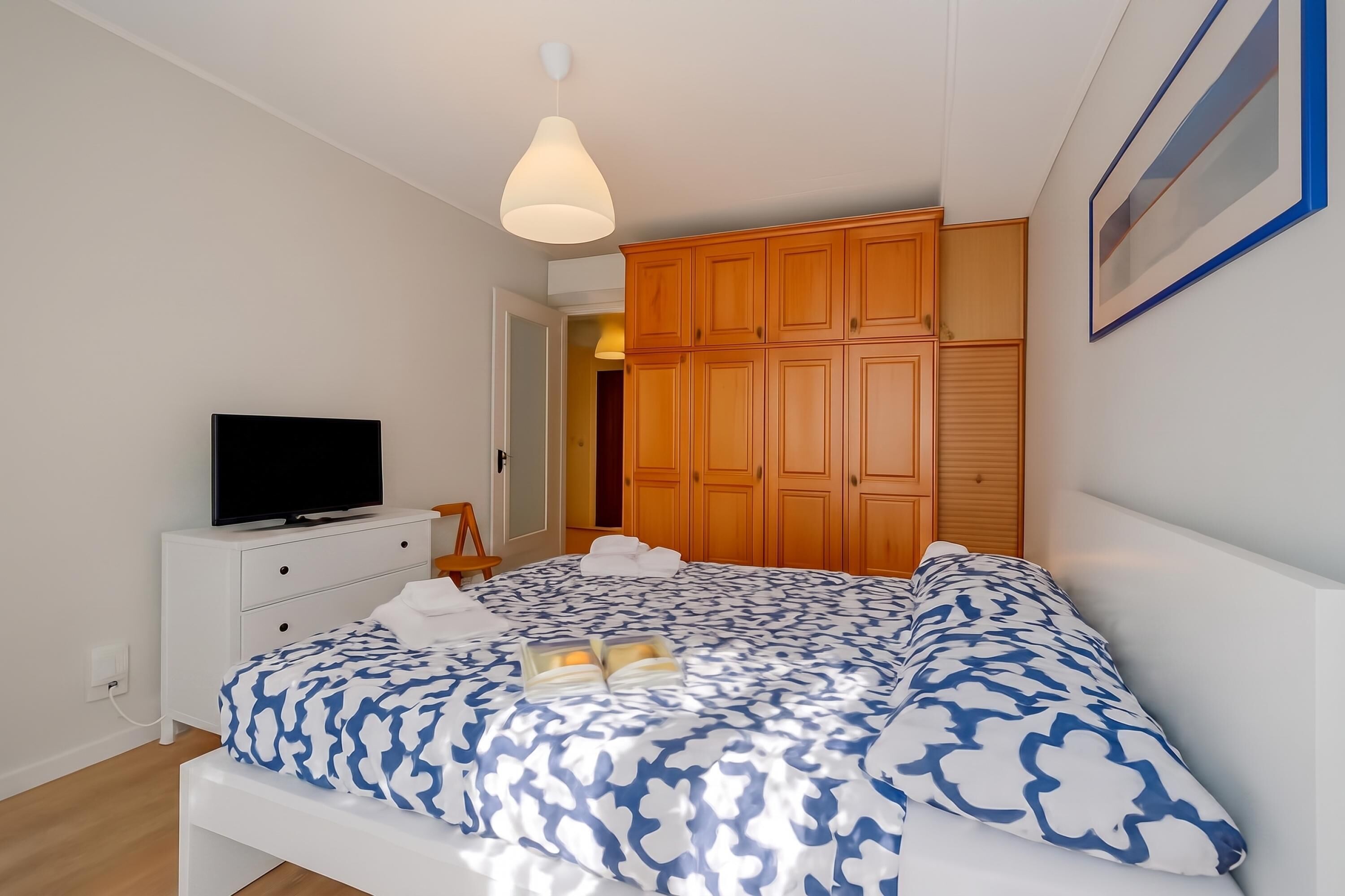 A bedroom with a bed, a television on a dresser, wooden wardrobe, and a pendant light.