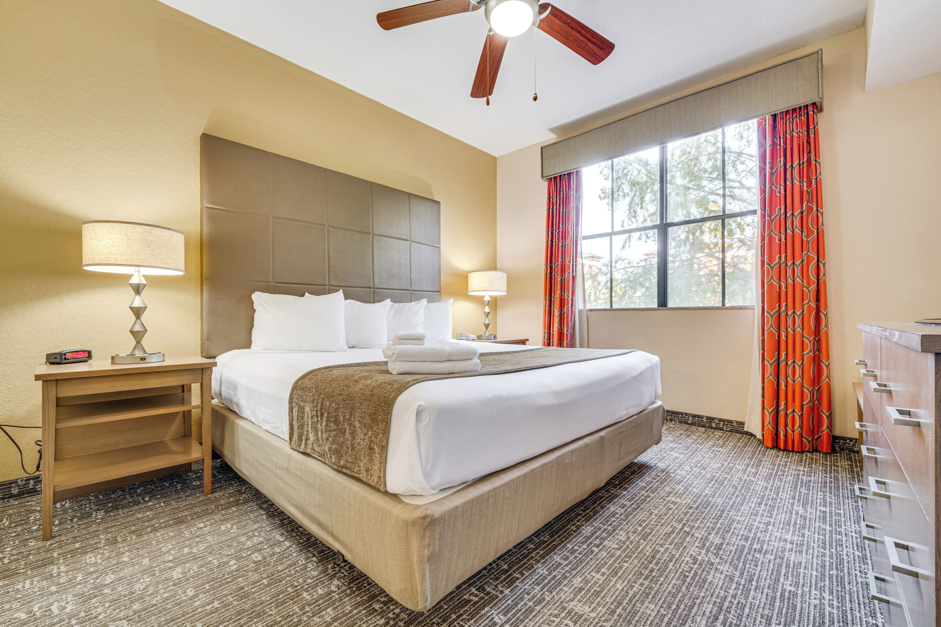 A hotel room with a large bed, two bedside tables with lamps, a ceiling fan, a window with curtains, and a dresser.