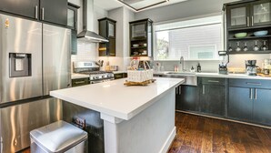 Une cuisine moderne dotée d’un îlot central, d’appareils électroménagers en acier inoxydable, de meubles de rangement aux couleurs sombres et d’une grande fenêtre qui laisse entrer la lumière naturelle.
