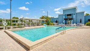 Una piscina con lettini e ombrelloni, circondata da un patio in mattoni e un edificio con facciata blu.