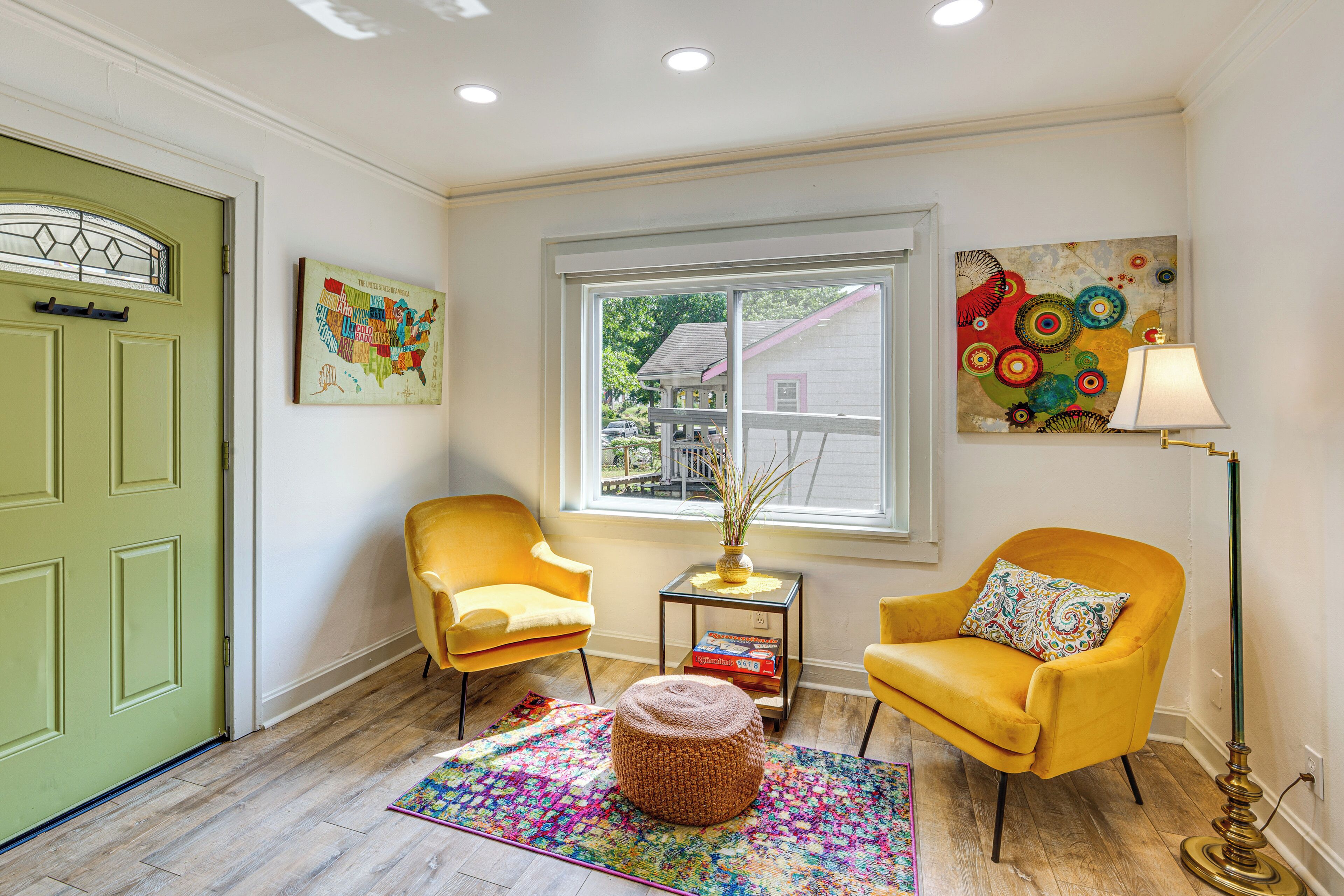 A room with a green door, two yellow armchairs, a colorful rug, a small table with a vase and books, and a window with a view of a house.