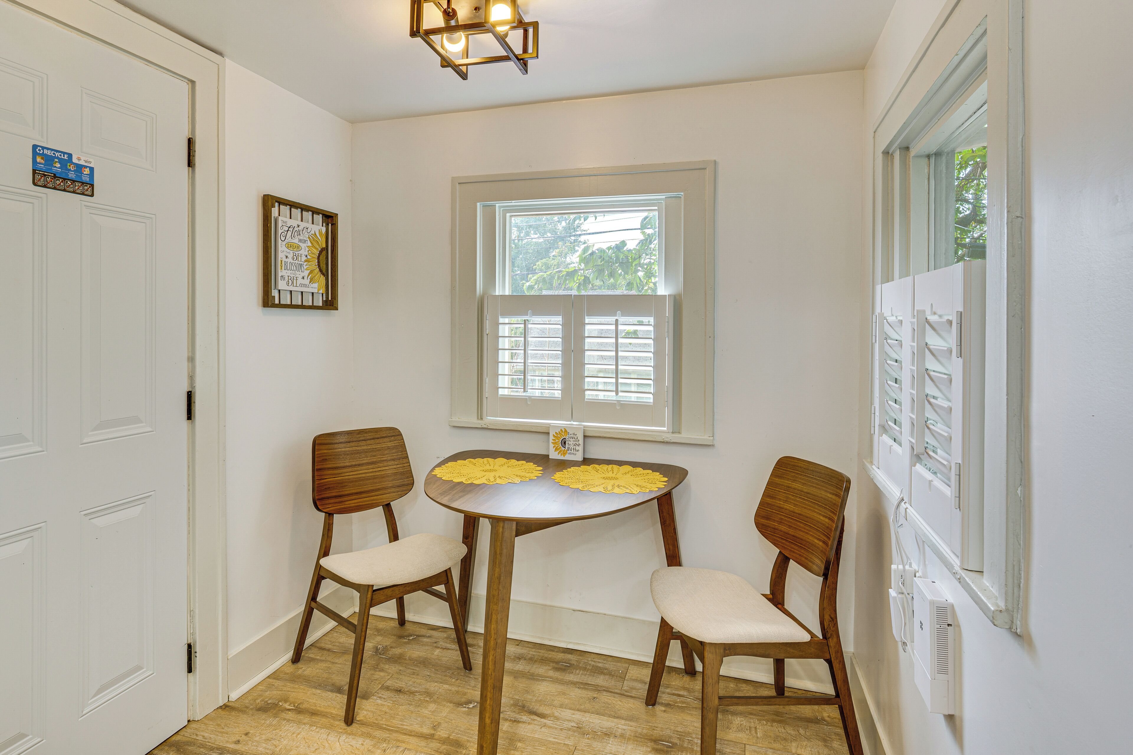 Una habitación con una mesa de comedor, dos sillas, una ventana con persianas y una lámpara en el techo.