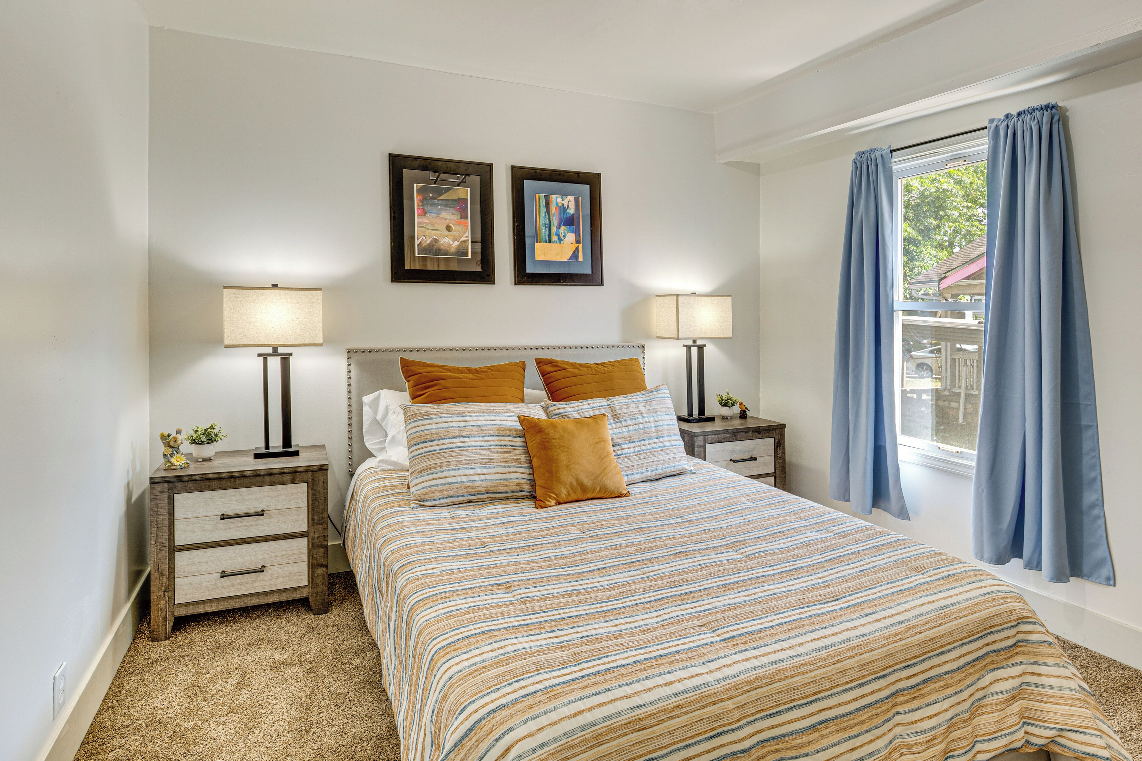 A bedroom with a bed, two bedside tables, a nightstand, a window with curtains, and two framed pictures on the wall.