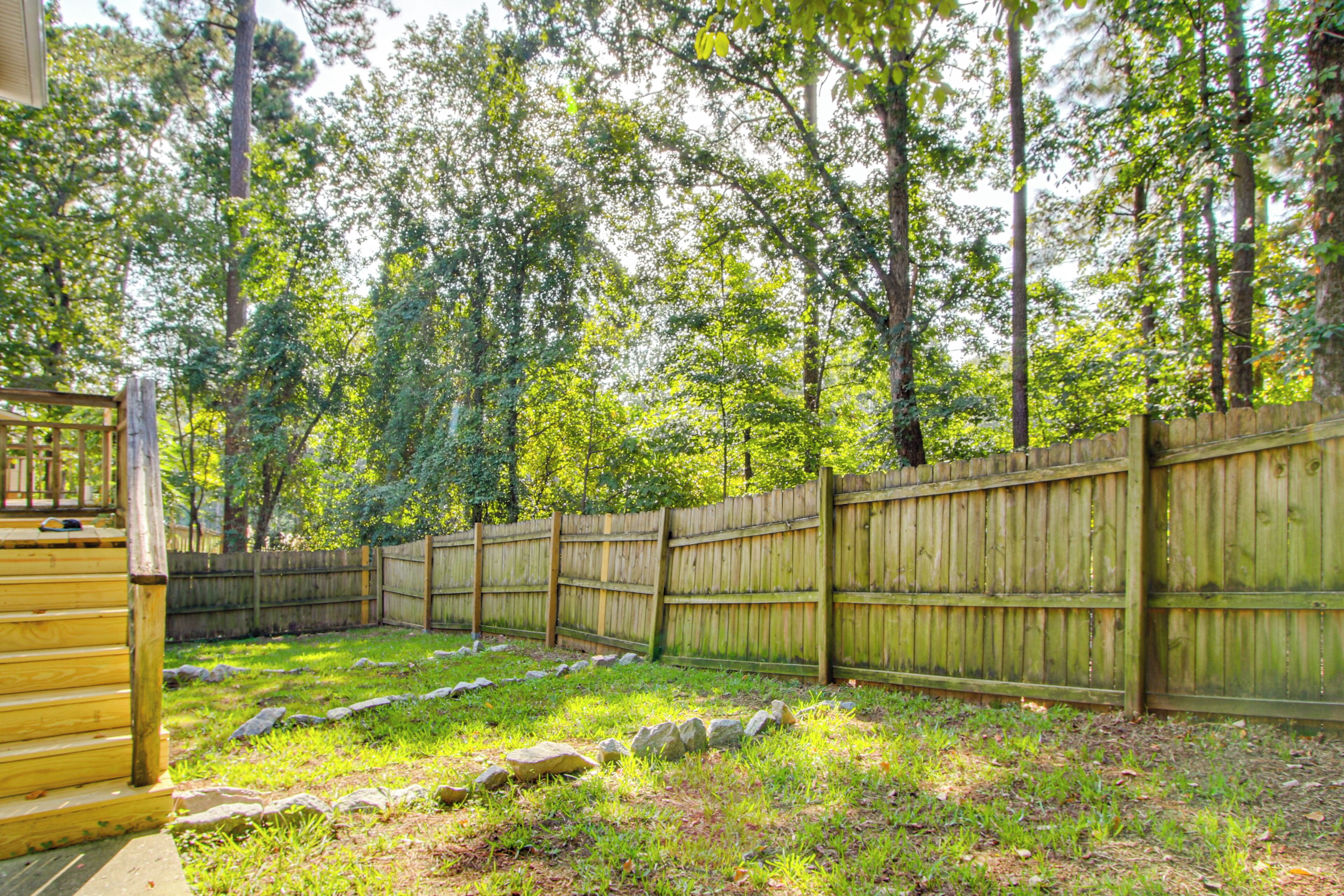 A fenced backyard with a wooden deck, surrounded by trees and a grassy area with rocks.