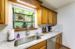 Una cocina con armarios de madera, un lavamanos de acero inoxidable, un ventanal con vistas a zonas verdes y una encimera con diversos objetos.