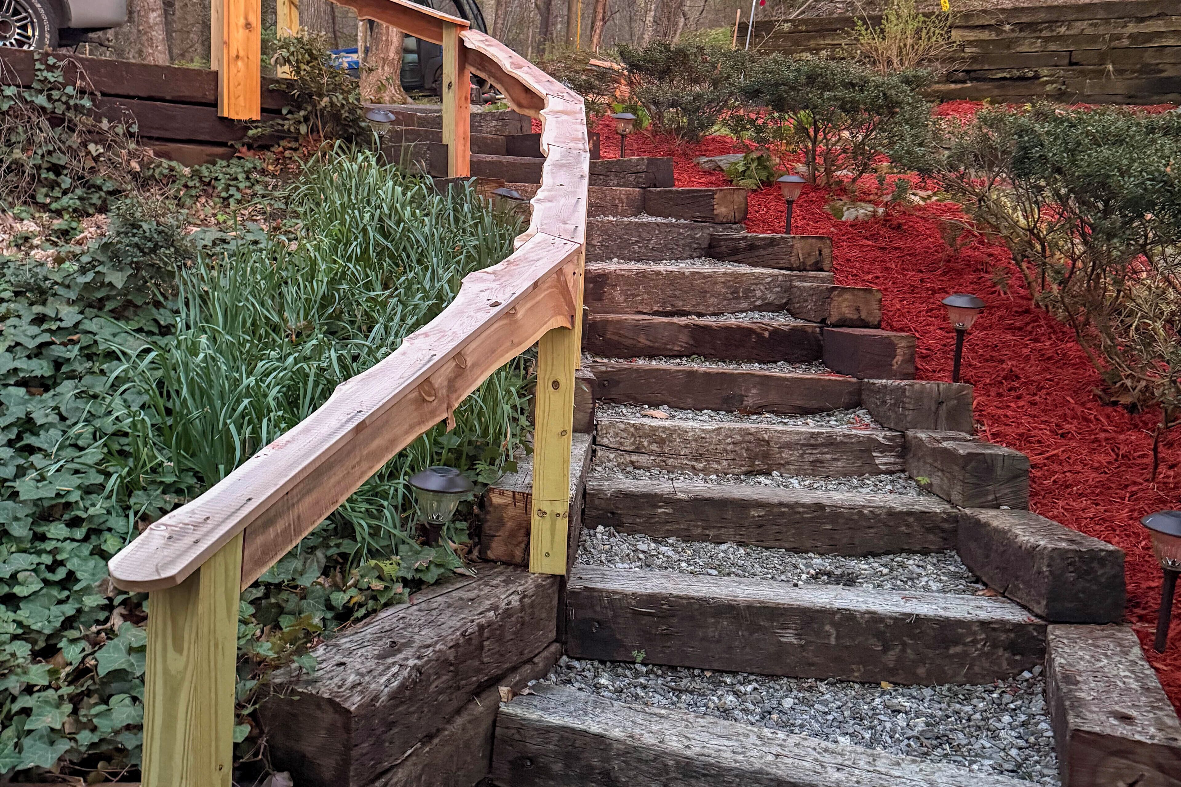 A wooden staircase with a curved handrail, surrounded by greenery and red mulch.
