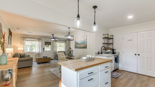 Un vaste espace de vie doté d’un plancher en bois, d’un îlot central avec des tiroirs et d’un coin repas comprenant une table et des chaises. On y trouve également une cuisine avec des armoires blanches et une porte double portant l’inscription « buanderie ».