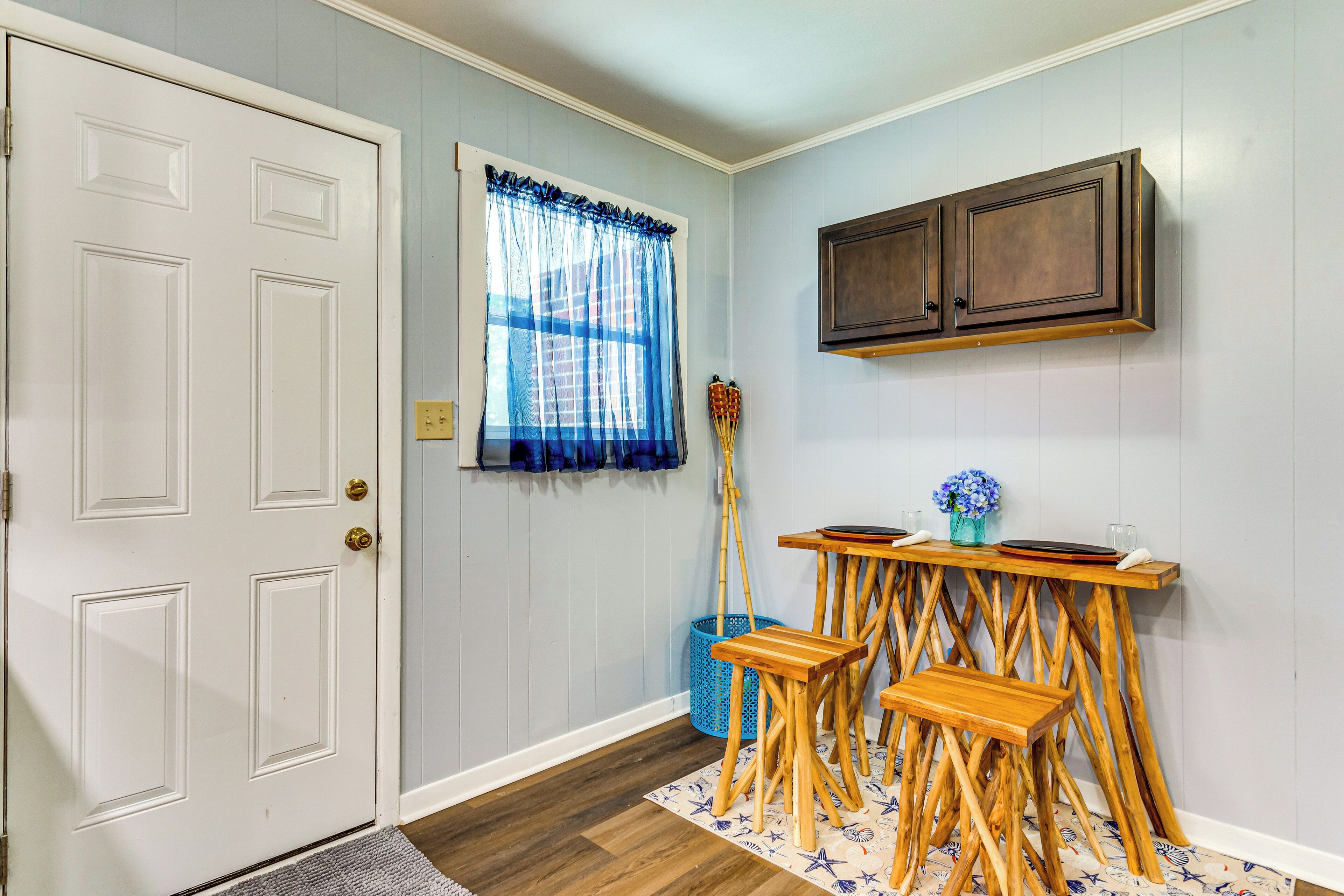 A room with a white door, a window with blue curtains, a wooden table with stools, and a dark wooden cabinet above the table.