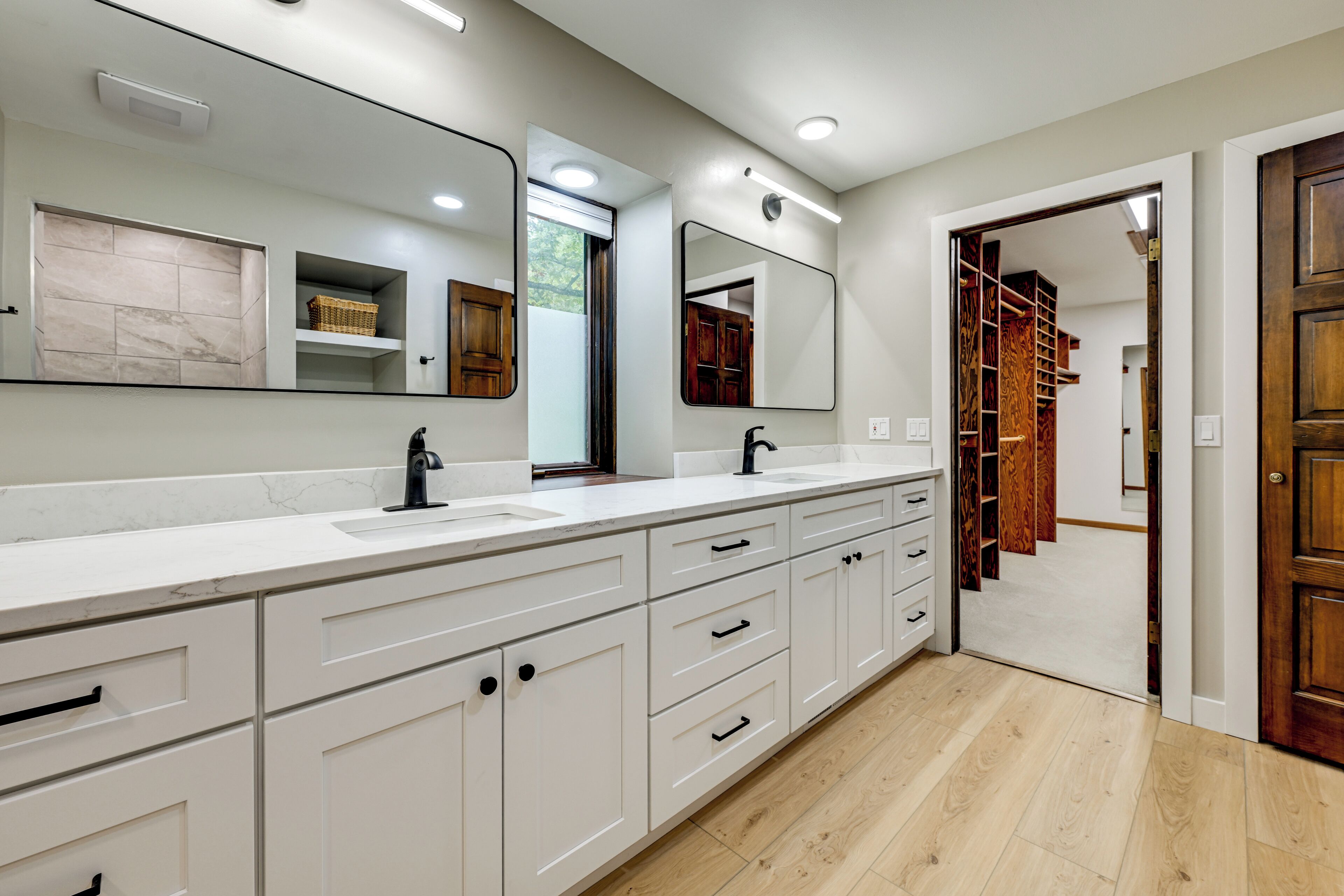 A modern bathroom with a large mirror, white cabinetry, and a wooden staircase leading to a closet.