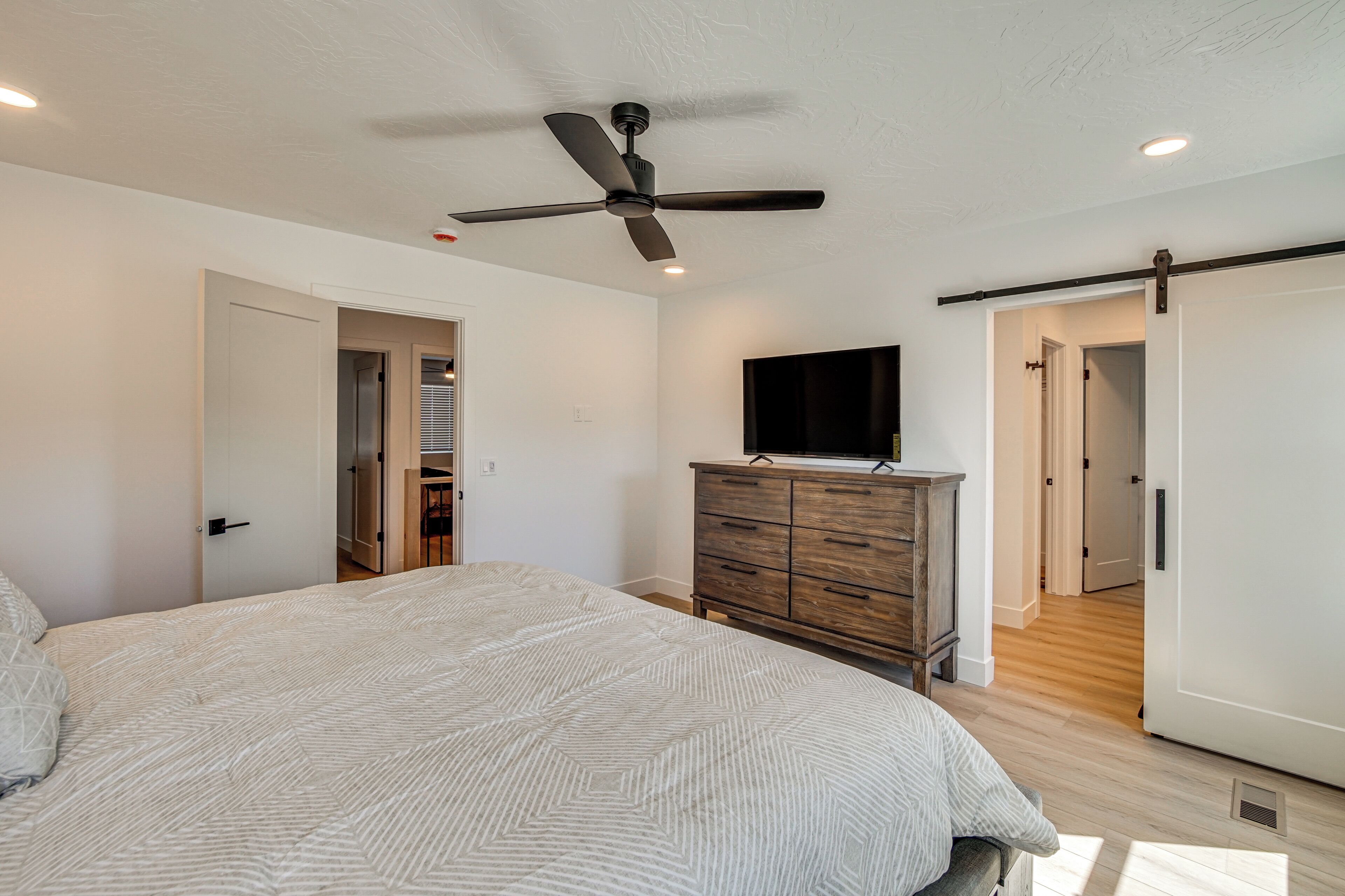 A bedroom with a bed, a ceiling fan, a wooden dresser, and a TV on the wall.