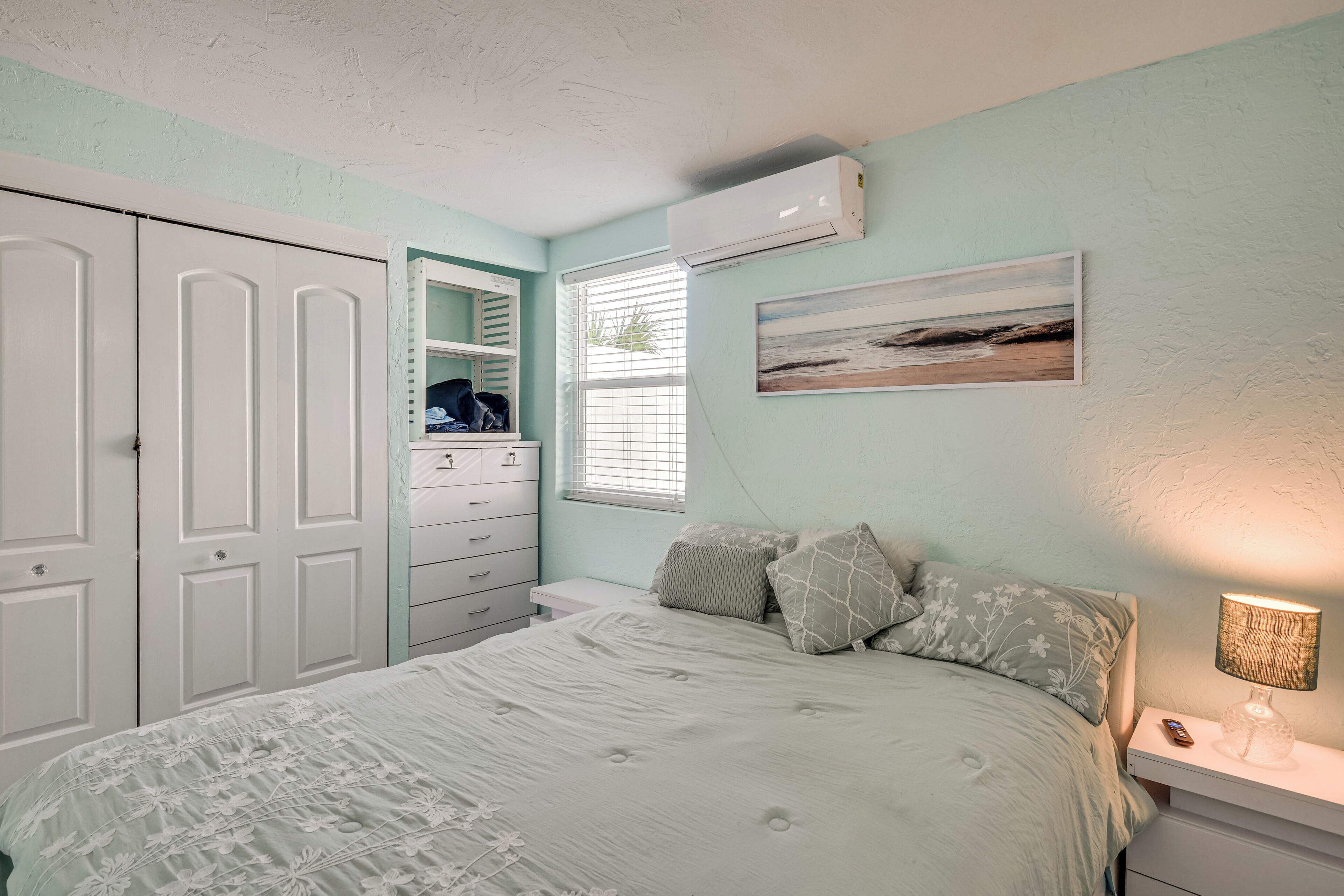 A bedroom with a bed, a nightstand, a dresser, an air conditioning unit, and a window with blinds.