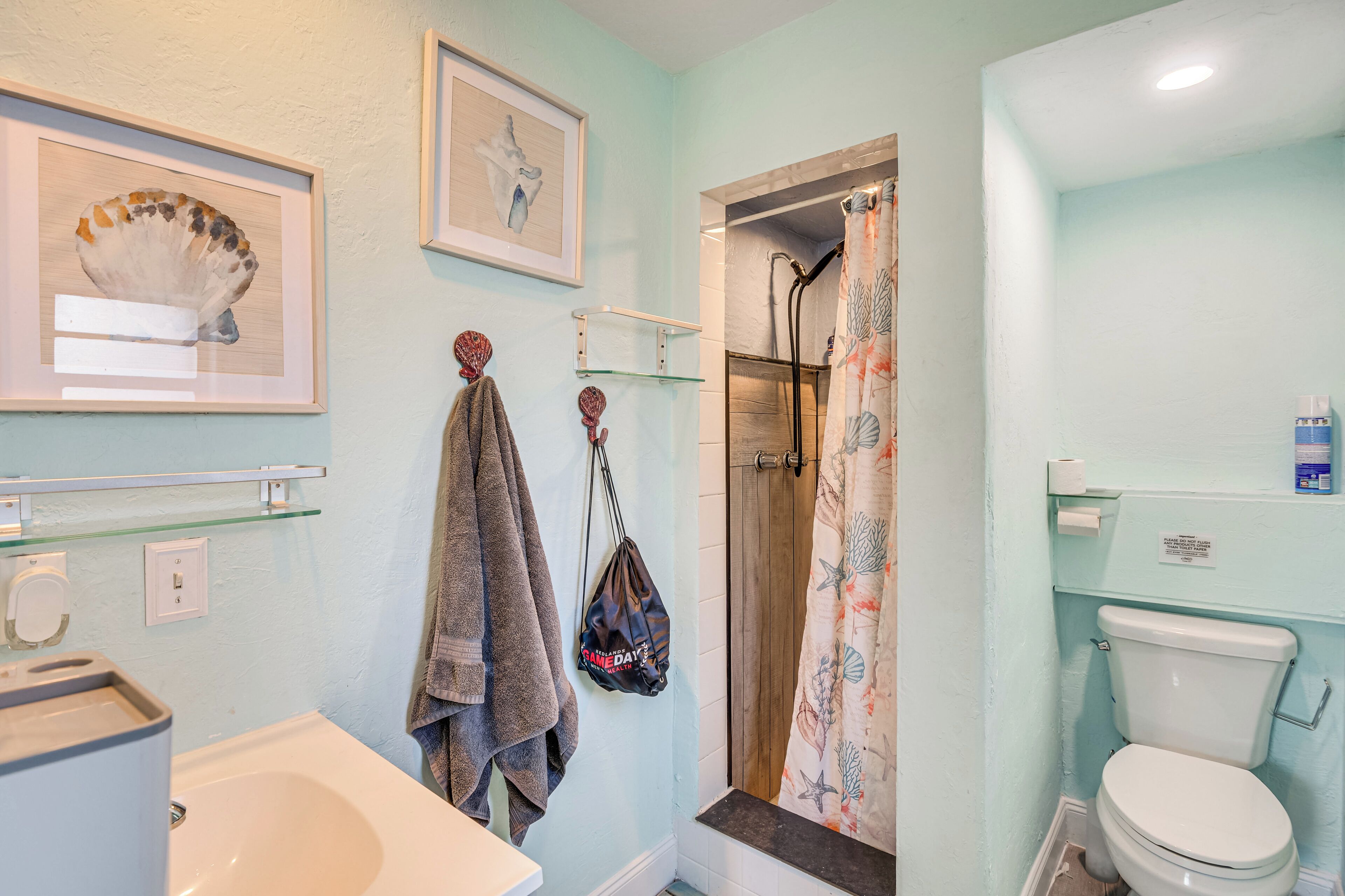 Et badeværelse med badekar, toilet, brusebad med blomstret bruseforhæng og billeder hængende på væggen.