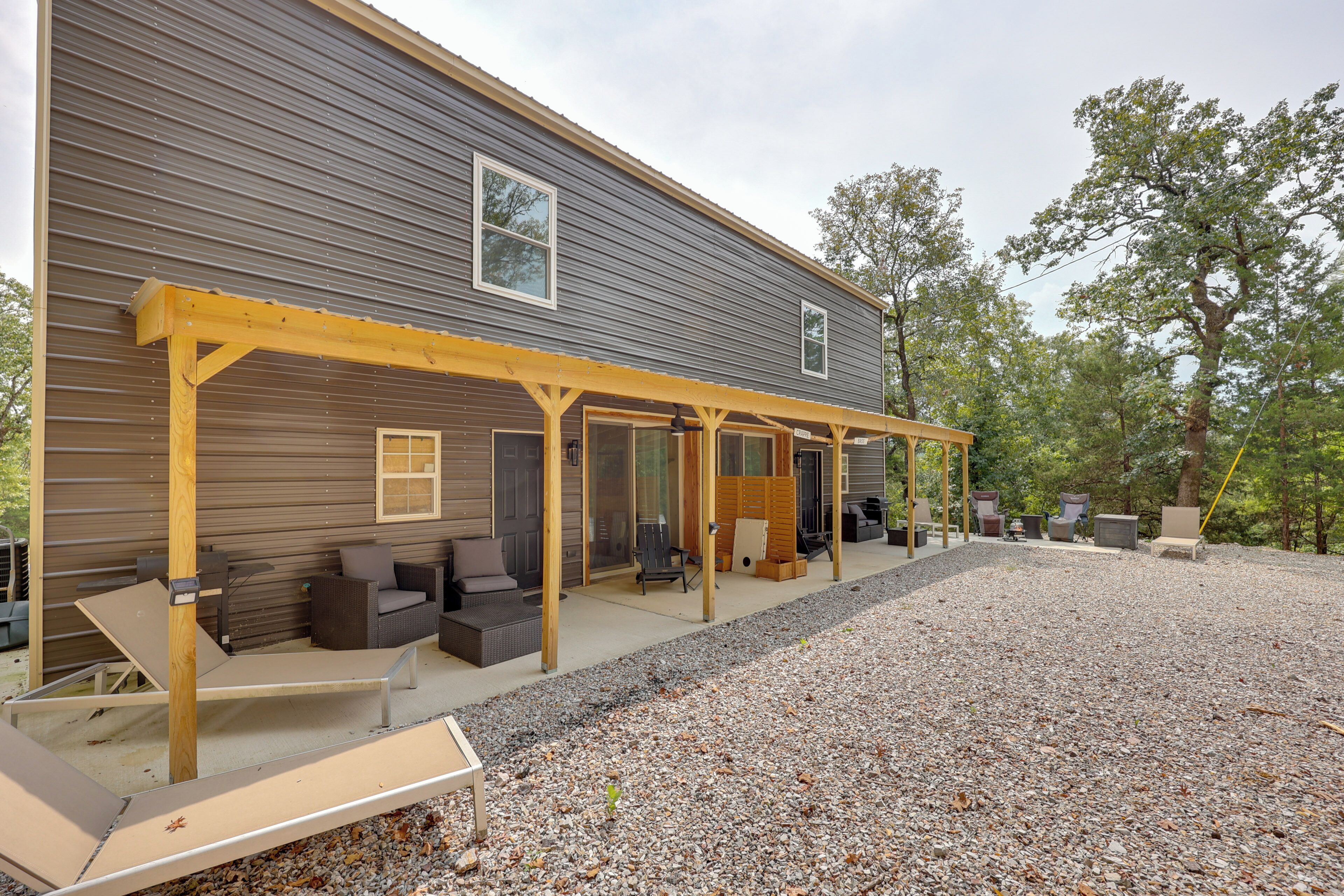A covered outdoor seating area with multiple chairs and tables, a gravel driveway, and a large building in the background.