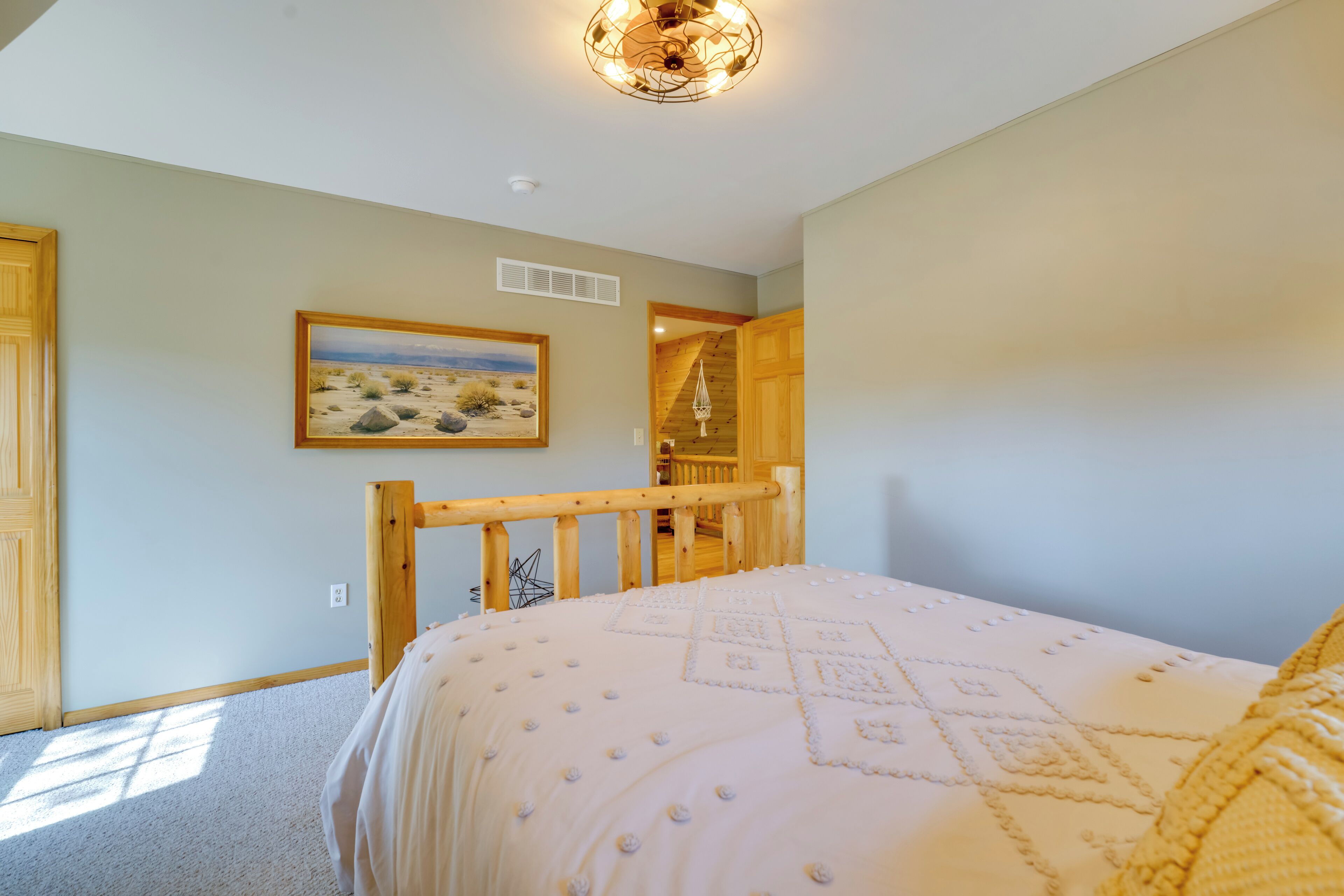A bedroom with a bed, a wooden headboard, a ceiling light fixture, a painting on the wall, and a door leading to another room.