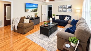 A living room with hardwood flooring, a ceiling fan, a flat-screen TV, a dark sofa, a brown armchair, and a coffee table with decorative items.