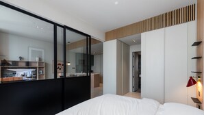 A modern hotel room with a large bed, a mirror, a wardrobe, and a kitchenette visible through a glass partition.