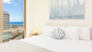 Une chambre d’hôtel avec une grande fenêtre, un lit recouvert de draps blancs, un coussin décoratif en forme de coquillage et un tableau accroché au mur.