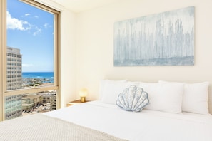 Une chambre d’hôtel avec une grande fenêtre, un lit recouvert de draps blancs, un coussin décoratif en forme de coquillage et un tableau accroché au mur.