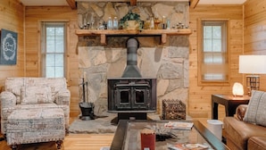 A cozy cabin interior with a stone fireplace, wooden walls, a patterned sofa, and a leather armchair.