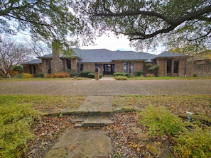 A brick house with a stone pathway leading to the front door, surrounded by trees and a gravel driveway. - The Castle / 9BR Retreat w/ Pool + Game Room (Garland)
