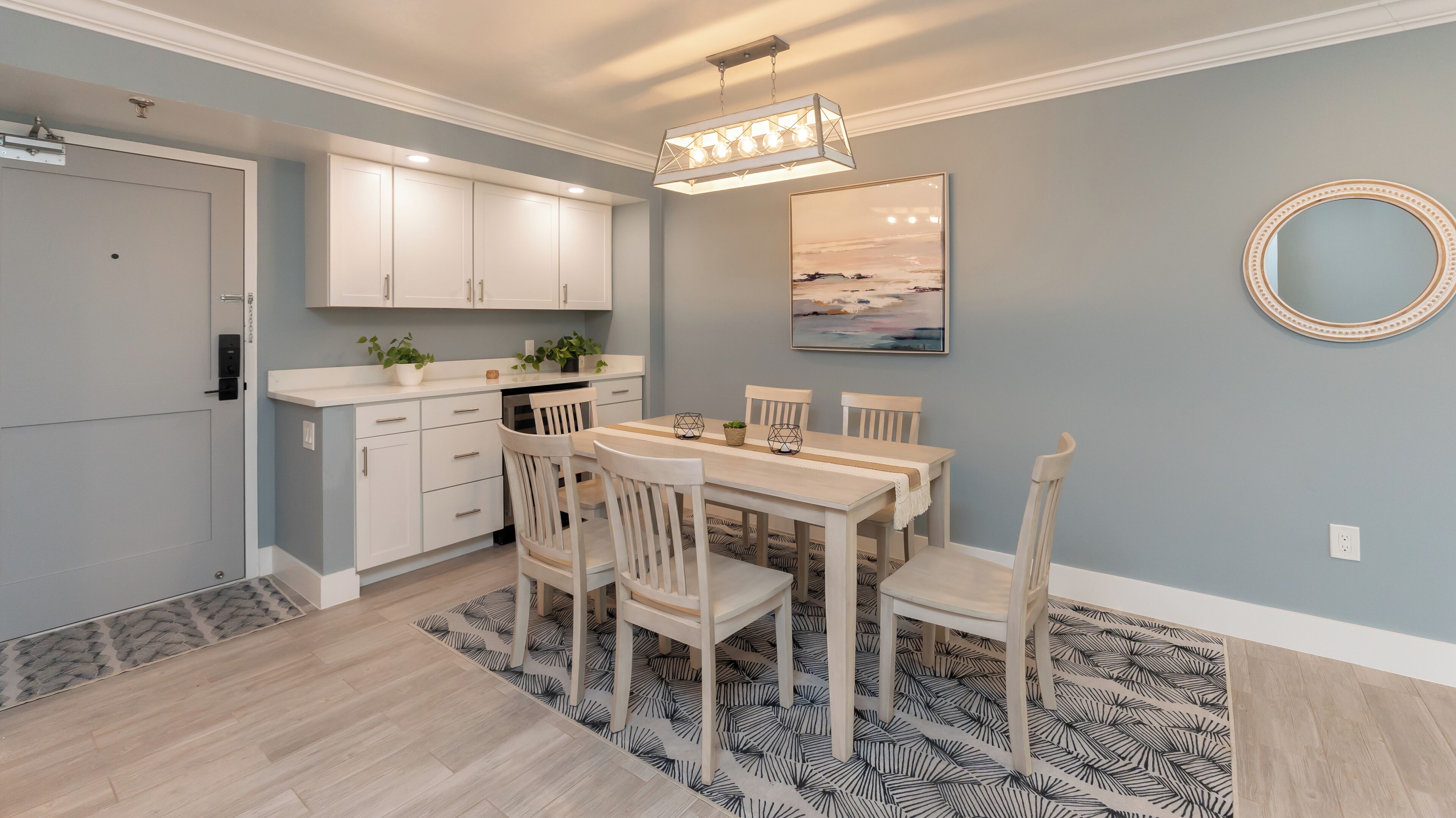 Une cuisine moderne avec des meubles blancs, un coin repas comprenant une table et des chaises en bois, un tapis à motifs et un luminaire au plafond.