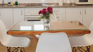 Una cocina moderna con gabinetes blancos, encimera de granito, mesa de vidrio, sillas blancas y un jarrón con flores.