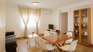 Un comedor con mesa de madera, sillas blancas, una mesita blanca, un televisor y un aparador con puertas de cristal.
