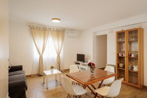 Un coin repas comprenant une table en bois, des chaises blanches, une petite table blanche, un téléviseur et un meuble avec des portes en verre.